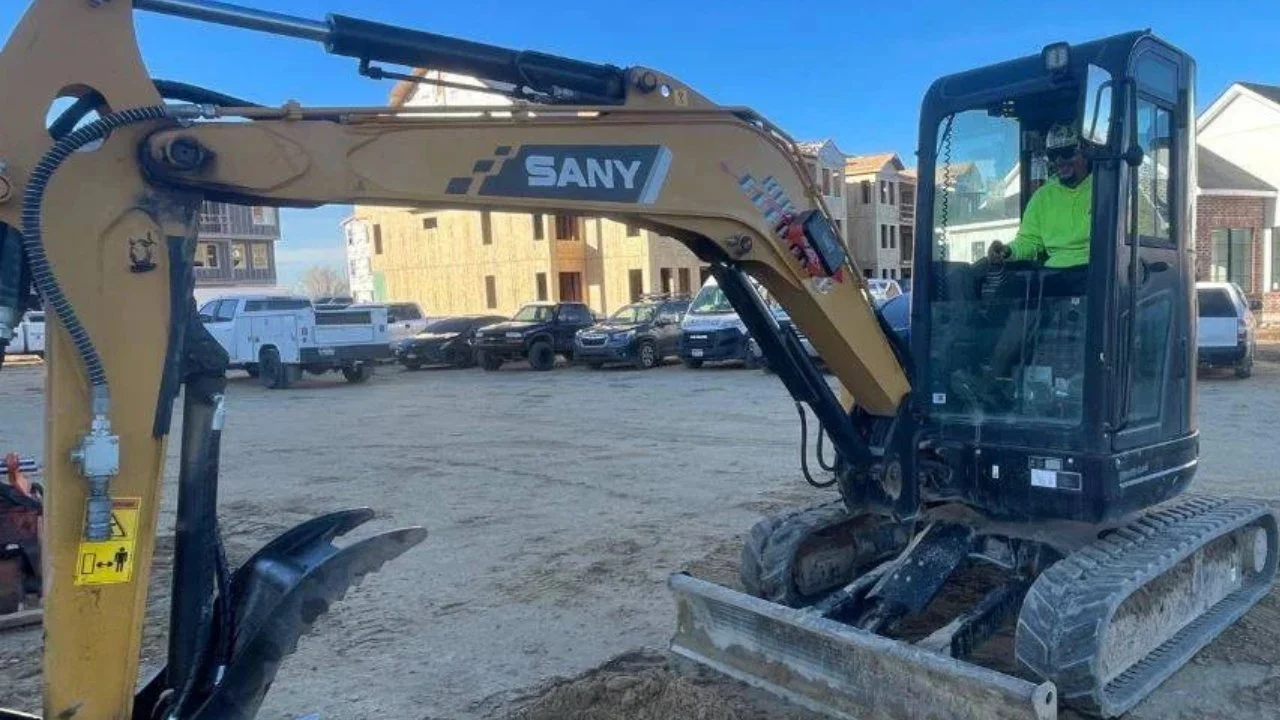 A construction worker operating a small SANY excavator on a dirt lot with residential houses and parked cars in the background.
