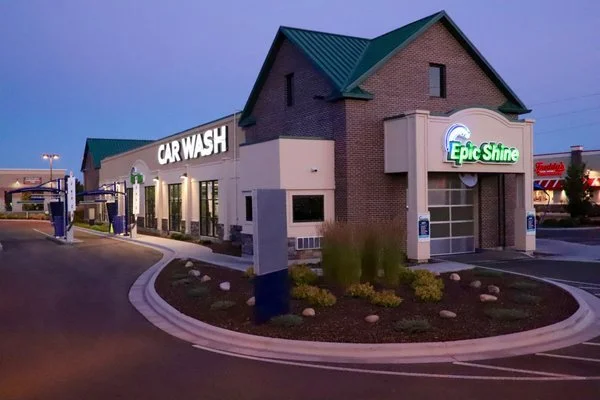 A car wash building with signs for 'Car Wash' and 'Epic Shine' during twilight, with a curved driveway and landscaped area in front.