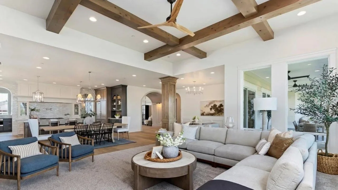 Spacious open-concept living and dining area with a white sectional sofa, wooden beams on the ceiling, and a dining table with chairs. Decor includes a large plant, table centerpiece, and artwork on the wall.