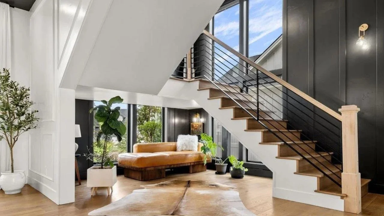 Interior view of a modern living room with a staircase, large windows, a tan leather sofa, potted plants, a cowhide rug, and black and white wall paneling.