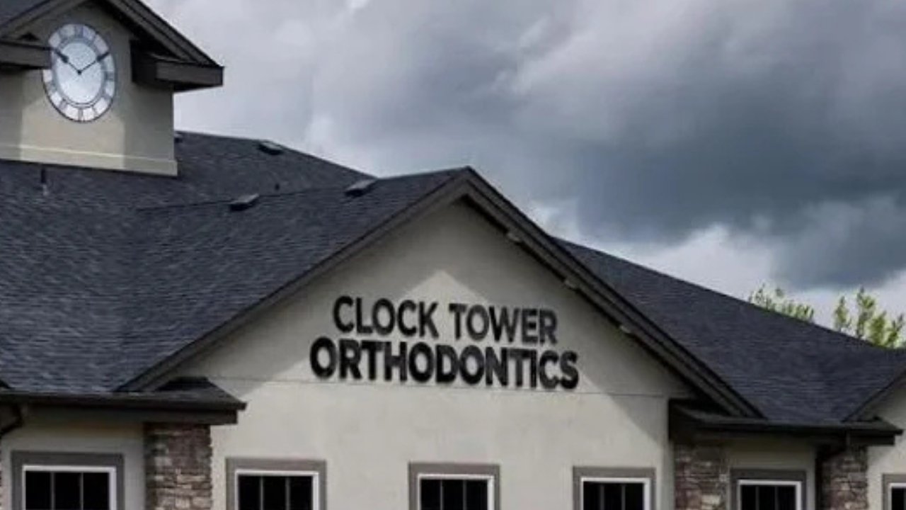 The exterior of a building with a sign that reads 'Clock Tower Orthodontics,' featuring a clock on the upper left side of the building and a dark cloudy sky above.