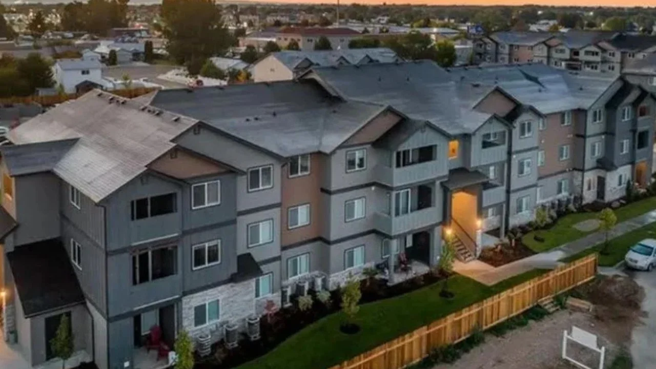 An aerial view of a multi-unit apartment complex with gray and beige exterior walls, small porches, green lawns, and a wooden fence. Some lights are on, and there are neighboring houses and streets in the background at sunset.