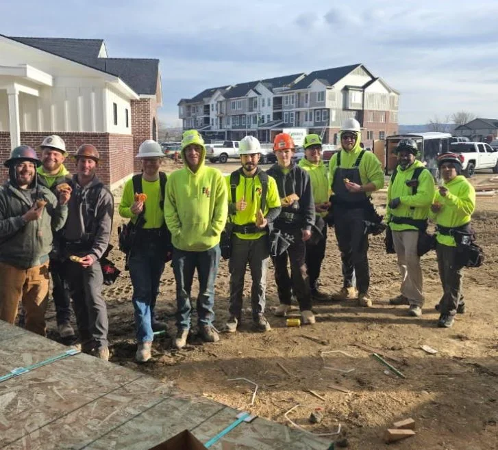 Group of construction workers standing on a dirt lot with houses and parked cars in the background. They are wearing safety helmets and high-visibility clothing.