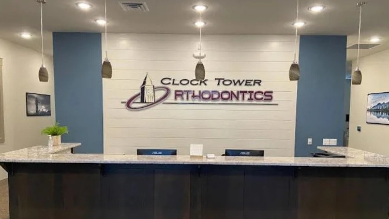 Reception area with a grey granite counter, a green plant, company logo on the wall reading 'Clock Tower Orthodontics', and hanging pendant lights.