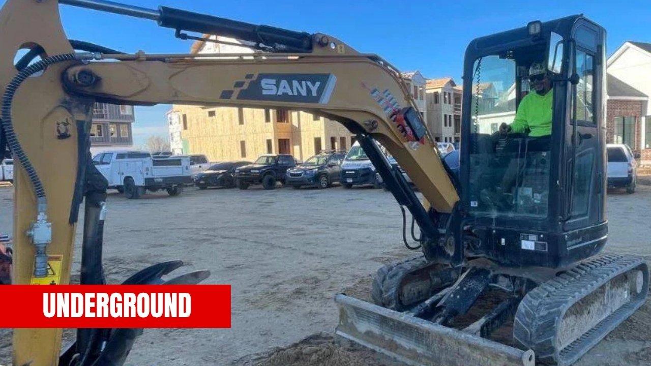A construction worker operating a yellow SANY excavator on a construction site with unfinished buildings and parked vehicles in the background.
