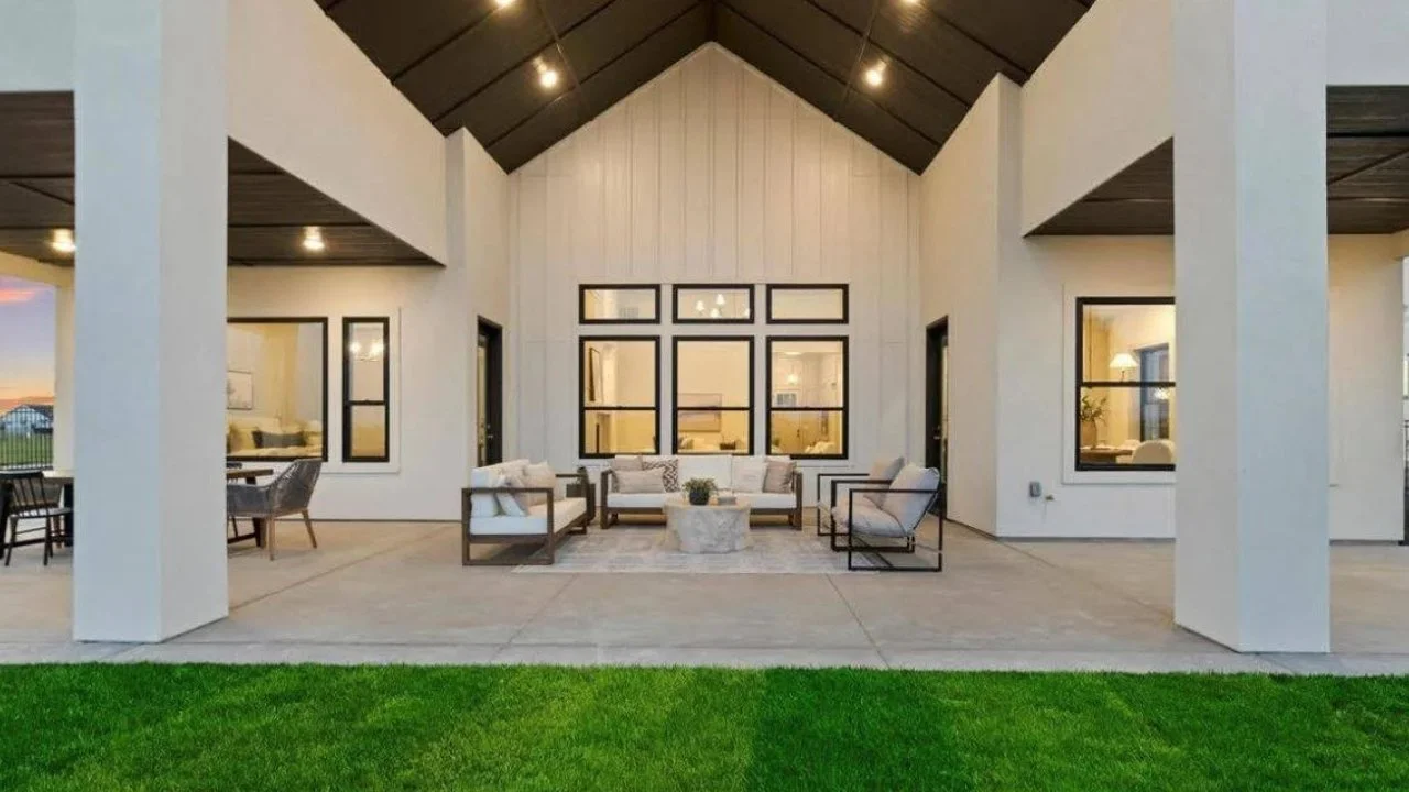 Exterior view of a modern porch with white walls, dark ceiling, and outdoor furniture including sofas and chairs around a small table, overlooking a green lawn.