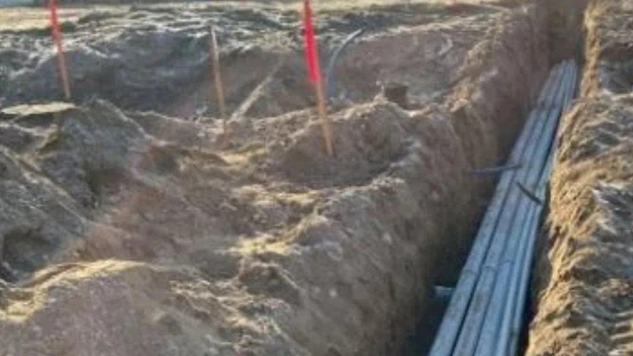 A construction excavation site with a deep trench and metal pipes laid along one side.