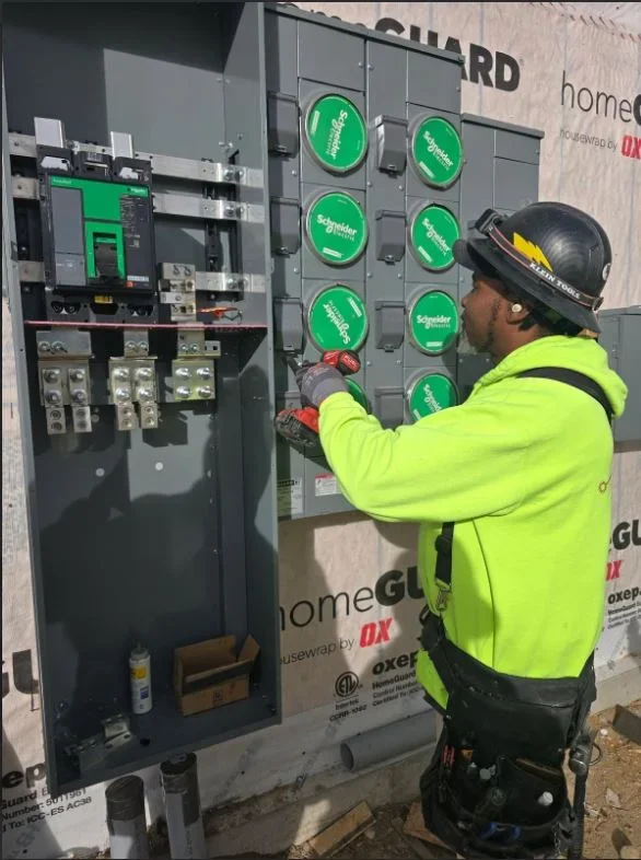 Electrician working on an electrical panel with circuit breakers and Schneider Electric equipment, wearing a hard hat and a neon yellow safety jacket.