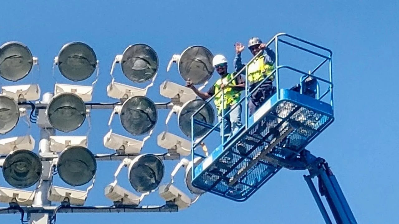 Two workers in safety gear and helmets standing on a blue lift platform next to a large group of floodlights mounted on a structure against a clear blue sky.