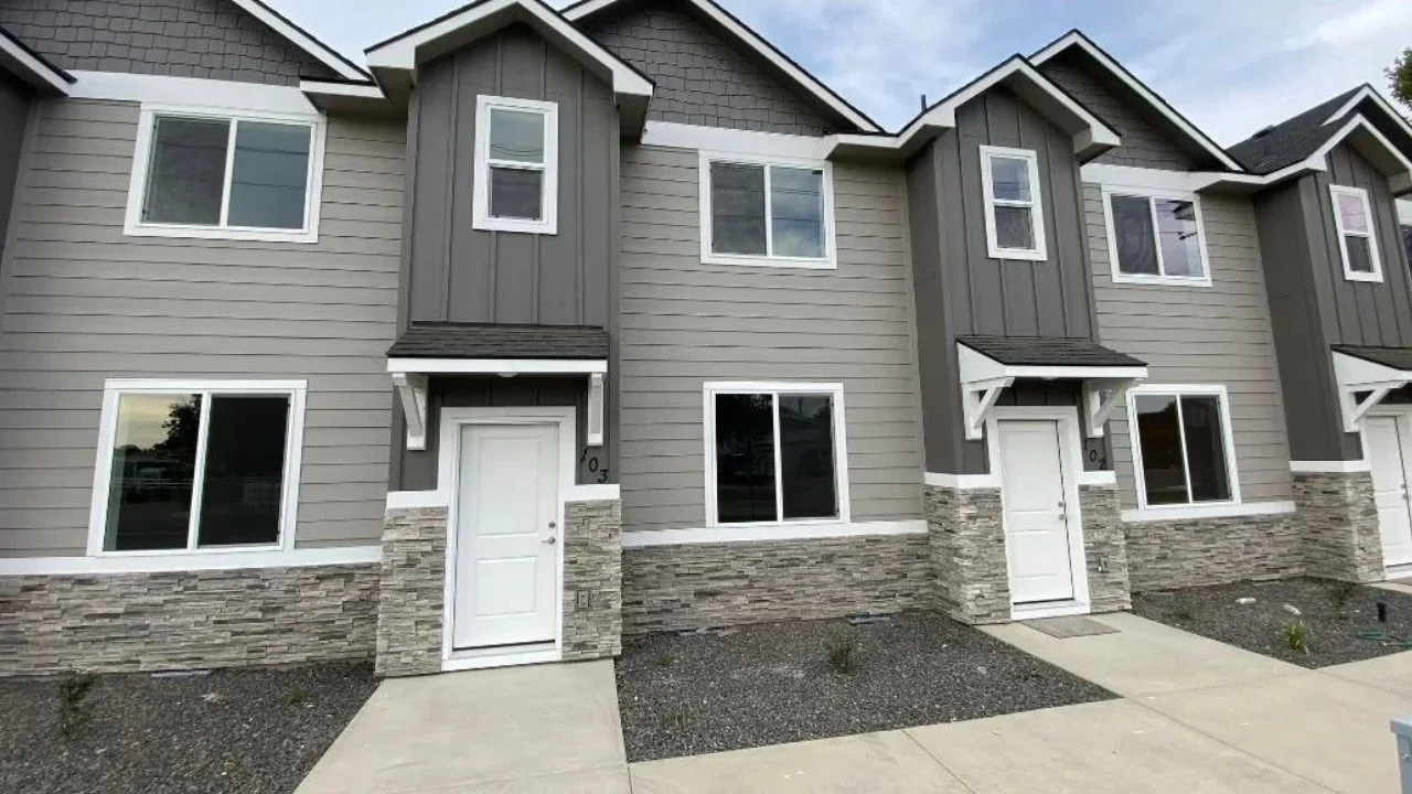 Front exterior view of modern multi-unit townhomes with gray siding, stone accents at the base, white doors, and multiple windows.