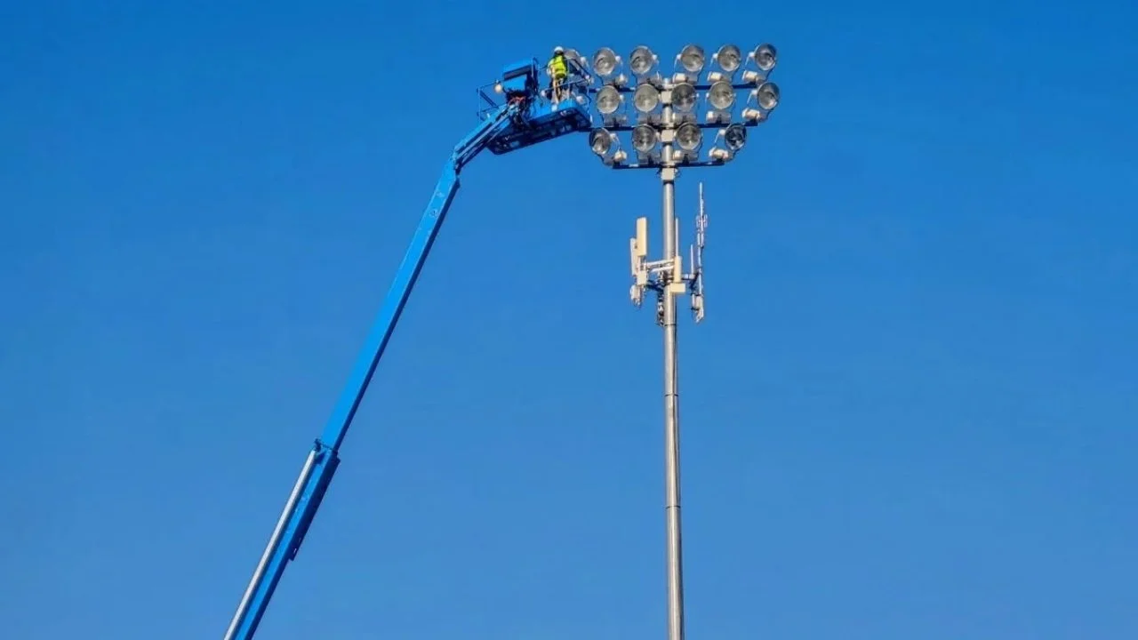 A person working on stadium floodlights on a high lift truck against a clear blue sky.