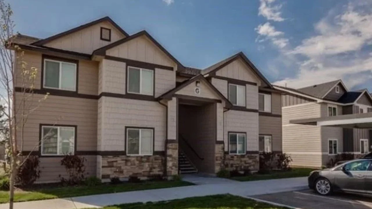 A two-story residential apartment building with beige and gray siding, stone accents at the base, multiple windows, and an entrance with stairs. Parked cars are visible in front.