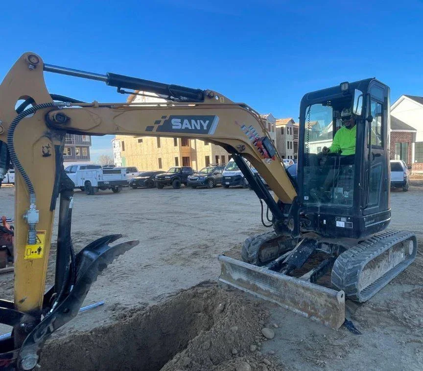 A small SANY excavator digging a trench at a construction site with residential homes in the background.