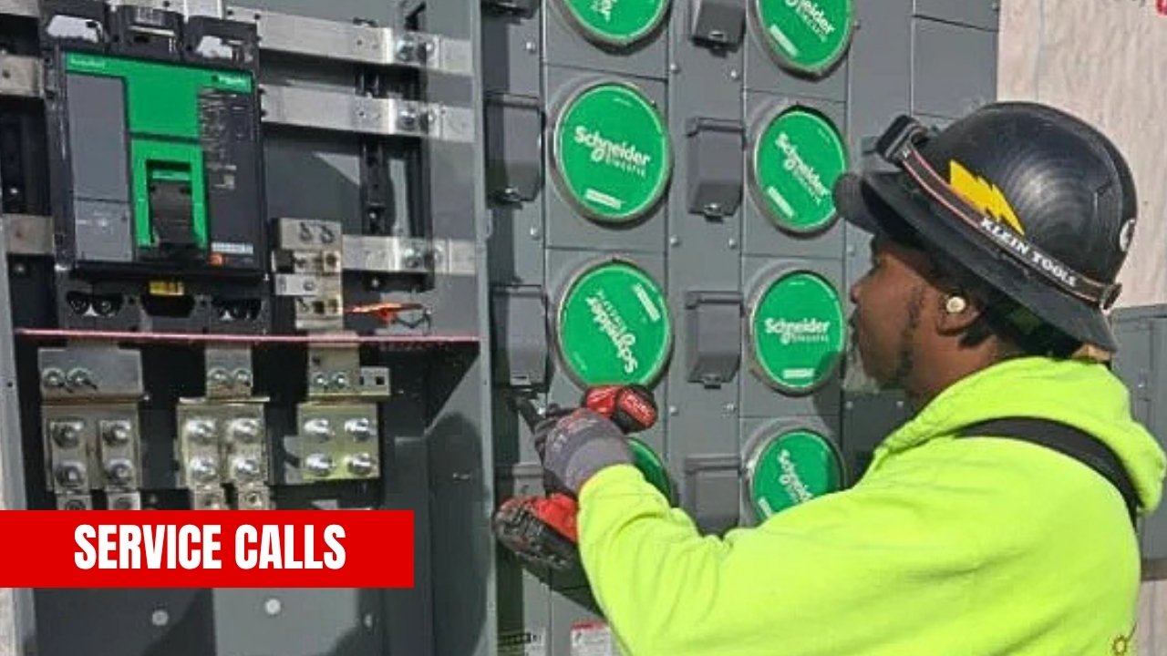 A worker wearing a black helmet with a headlamp and a yellow hoodie is inspecting or repairing an electrical panel with various circuit breakers and switches. There are green Schneider Electric circuit breakers on the panel. The text 'SERVICE CALLS' 