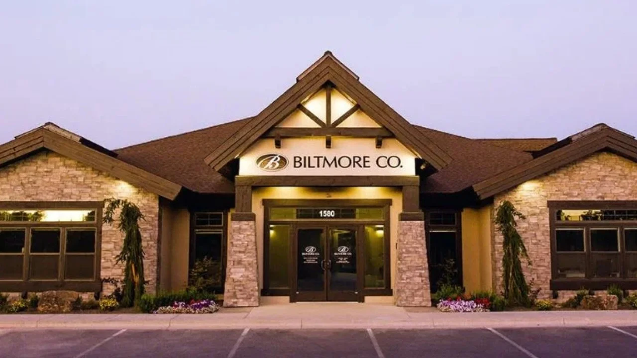 Exterior of a commercial building with a sign reading 'Biltmore Co.', stone brick facade, large windows, and flower beds in front.