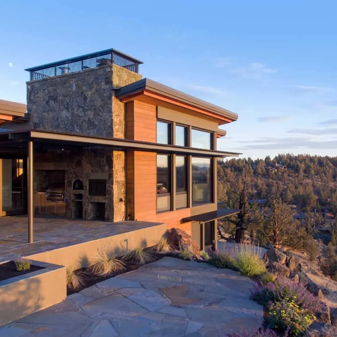 Modern house with stone and wood exterior on a hillside, featuring large windows and an outdoor patio with plants, overlooking a landscape with trees and hills.
