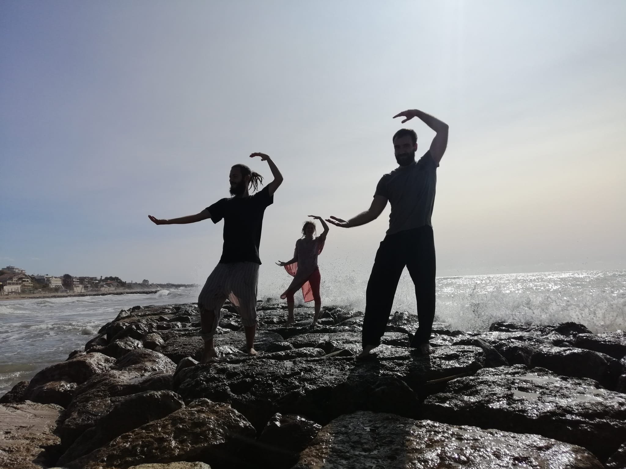 People standing on rocks near the ocean with waves crashing against the rocks, forming a silhouette against the bright sky.