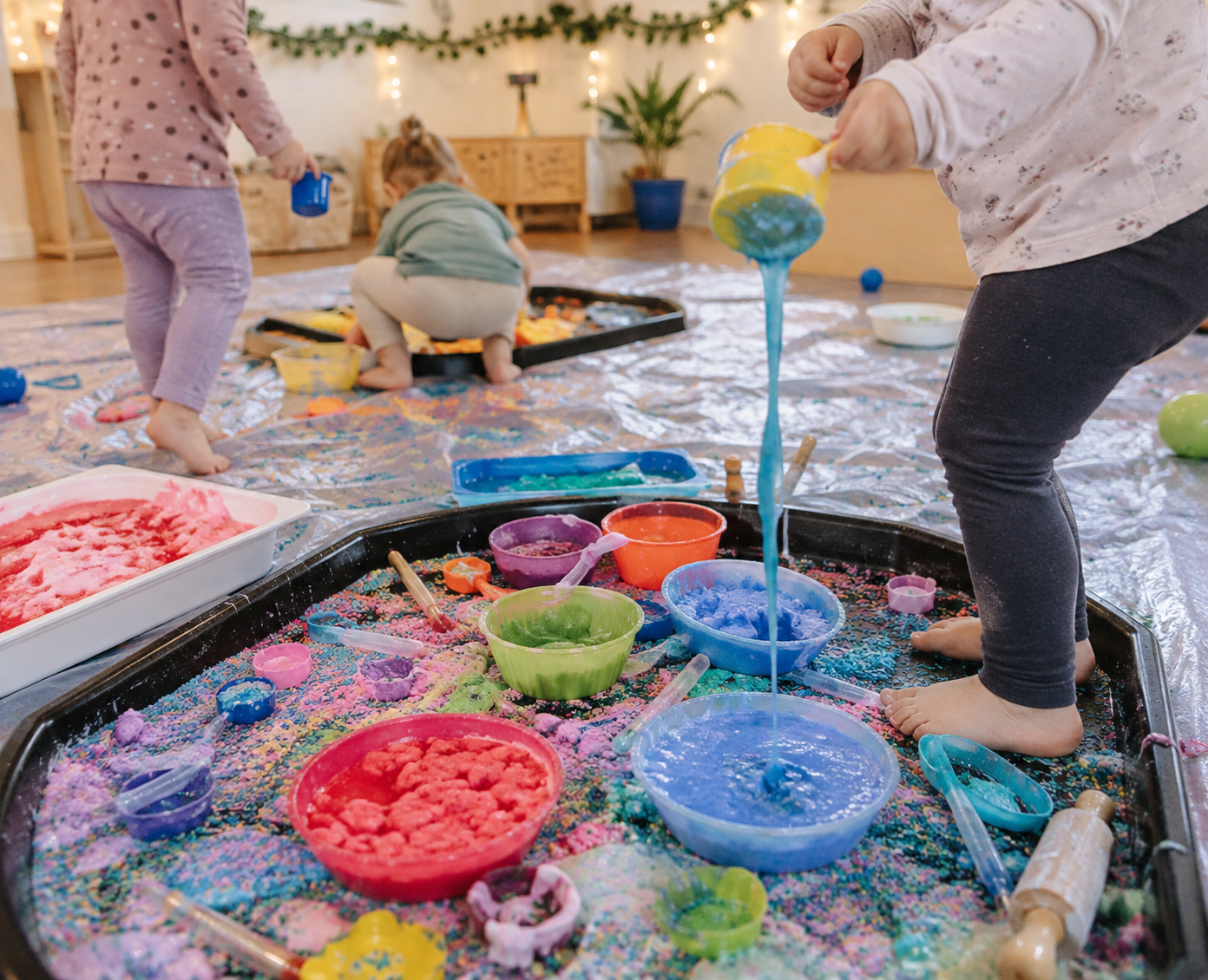 Kinder entdecken eine farbenfrohe Tiny Senses Messy Play Station mit bunten Materialien zum Giessen, Mischen, Fühlen und Experimentieren.