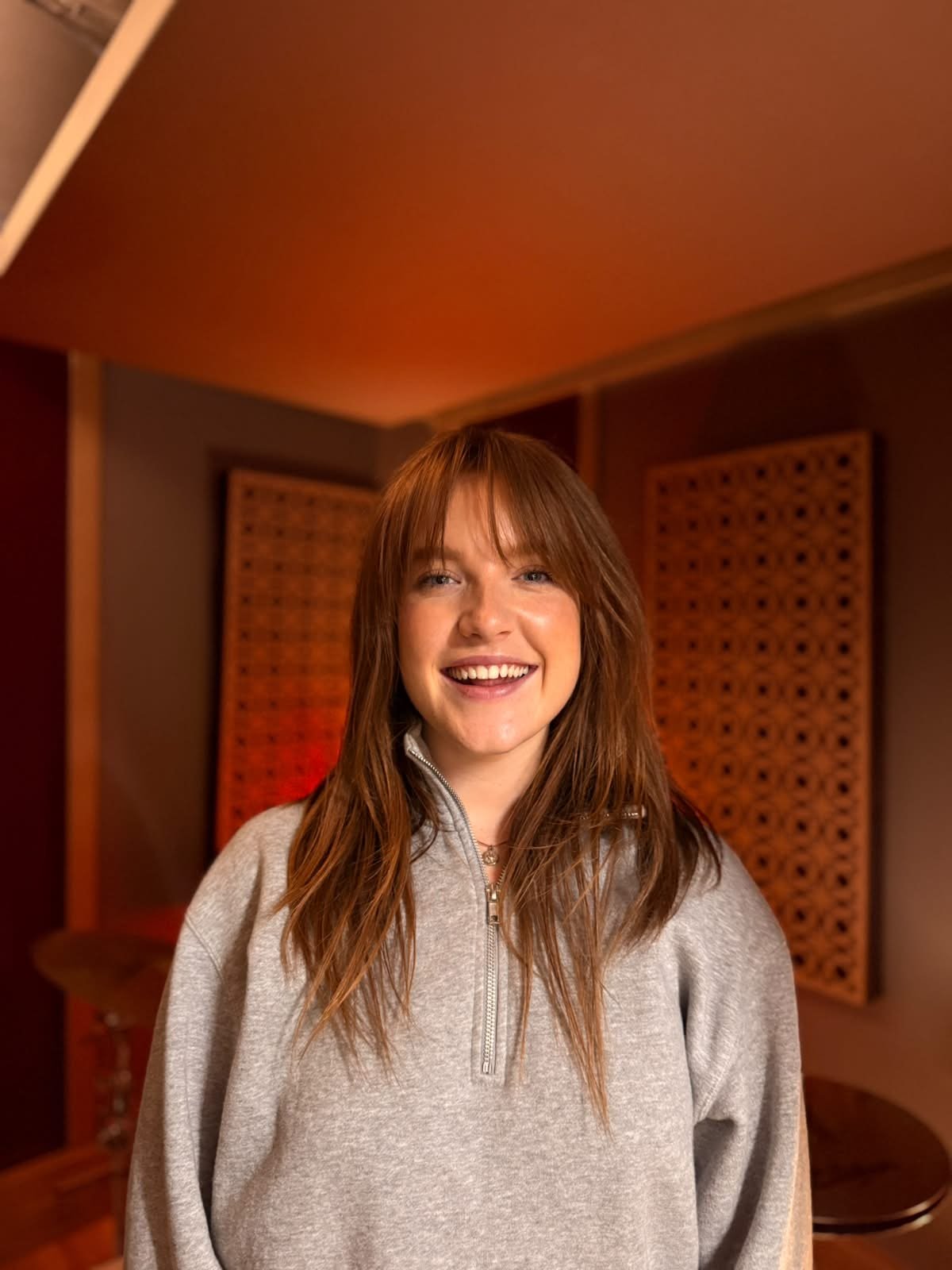A young woman with long reddish hair, smiling, wearing a gray zip-up hoodie, standing in a room with acoustic panels on the walls.