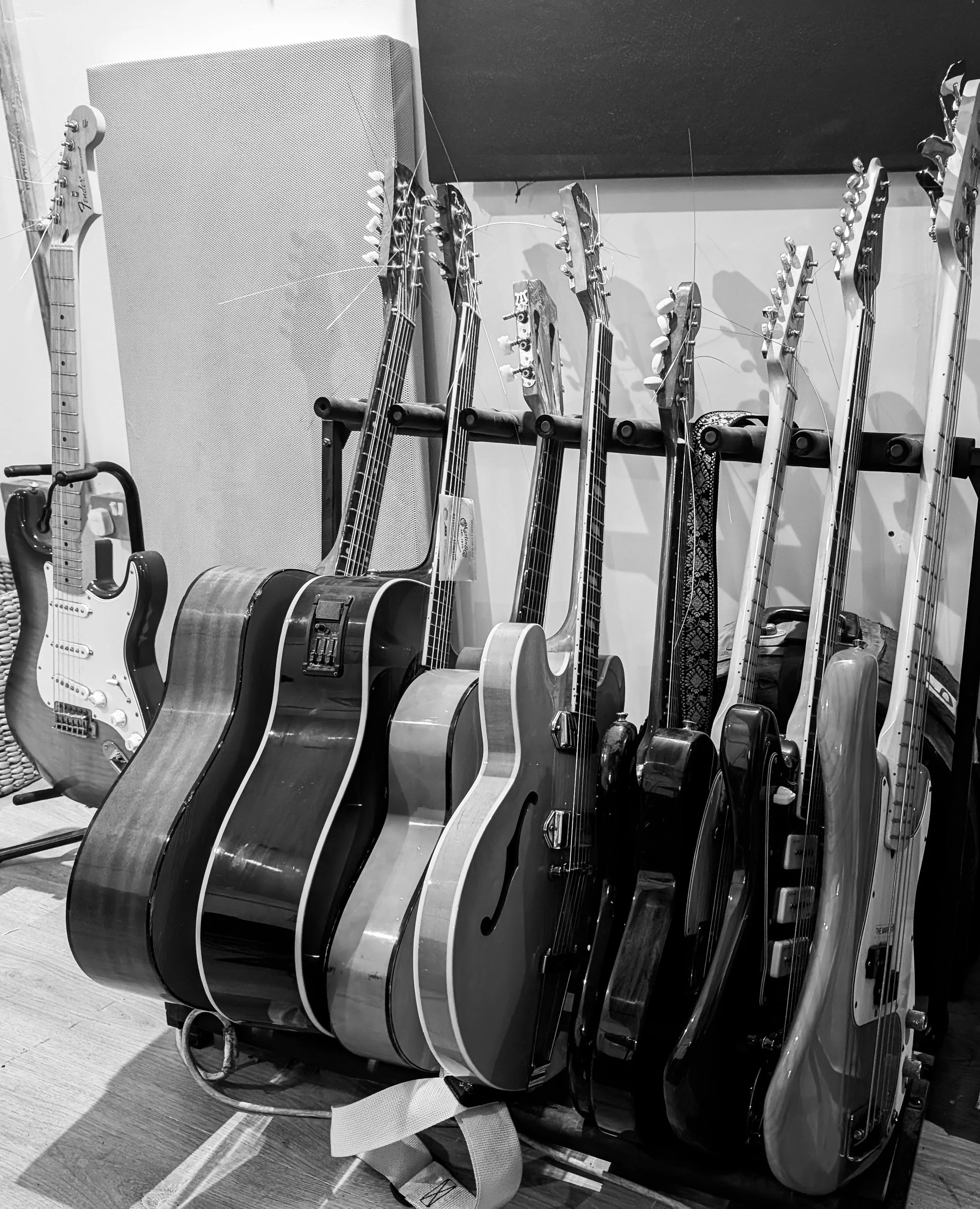 A row of guitars of various styles, including acoustic and electric, standing on a rack in a music store or studio, with some guitar straps visible.