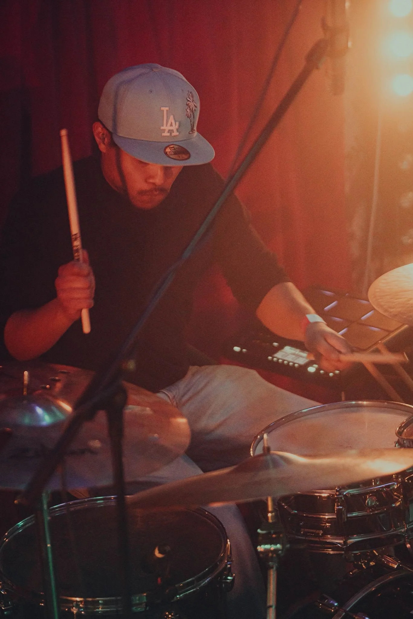 A man wearing a gray Los Angeles Dodgers cap playing a drum set in a dimly lit room.