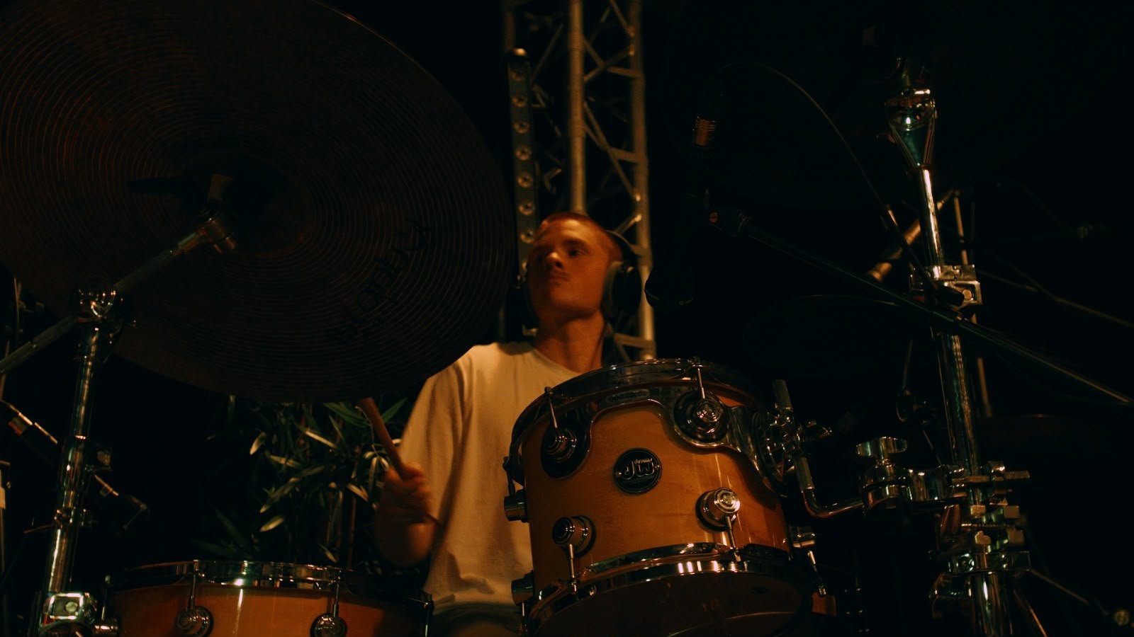 A young man wearing headphones playing the drums on stage with a large cymbal and drum set under stage lighting.
