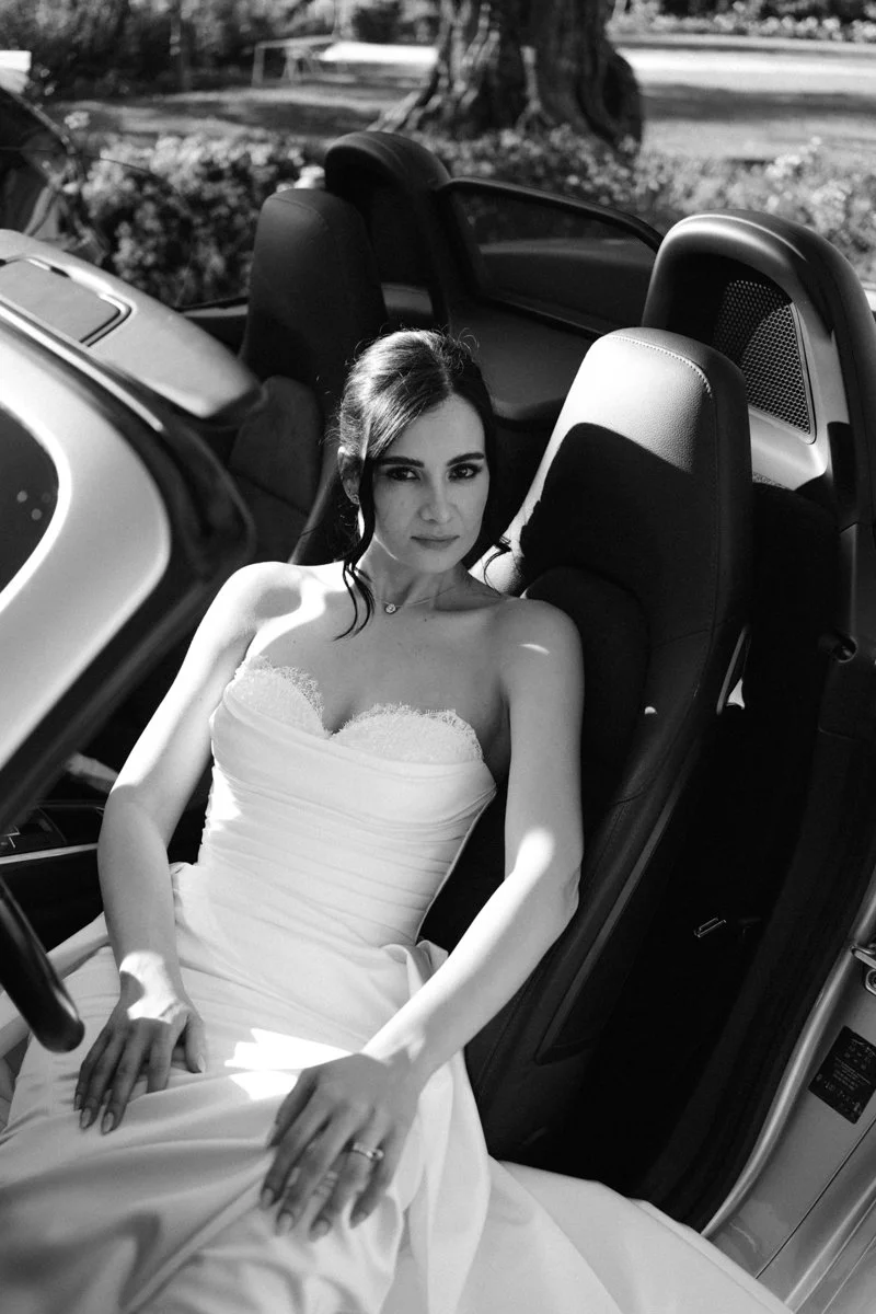 A woman in a wedding dress sitting in the driver's seat of a convertible car.