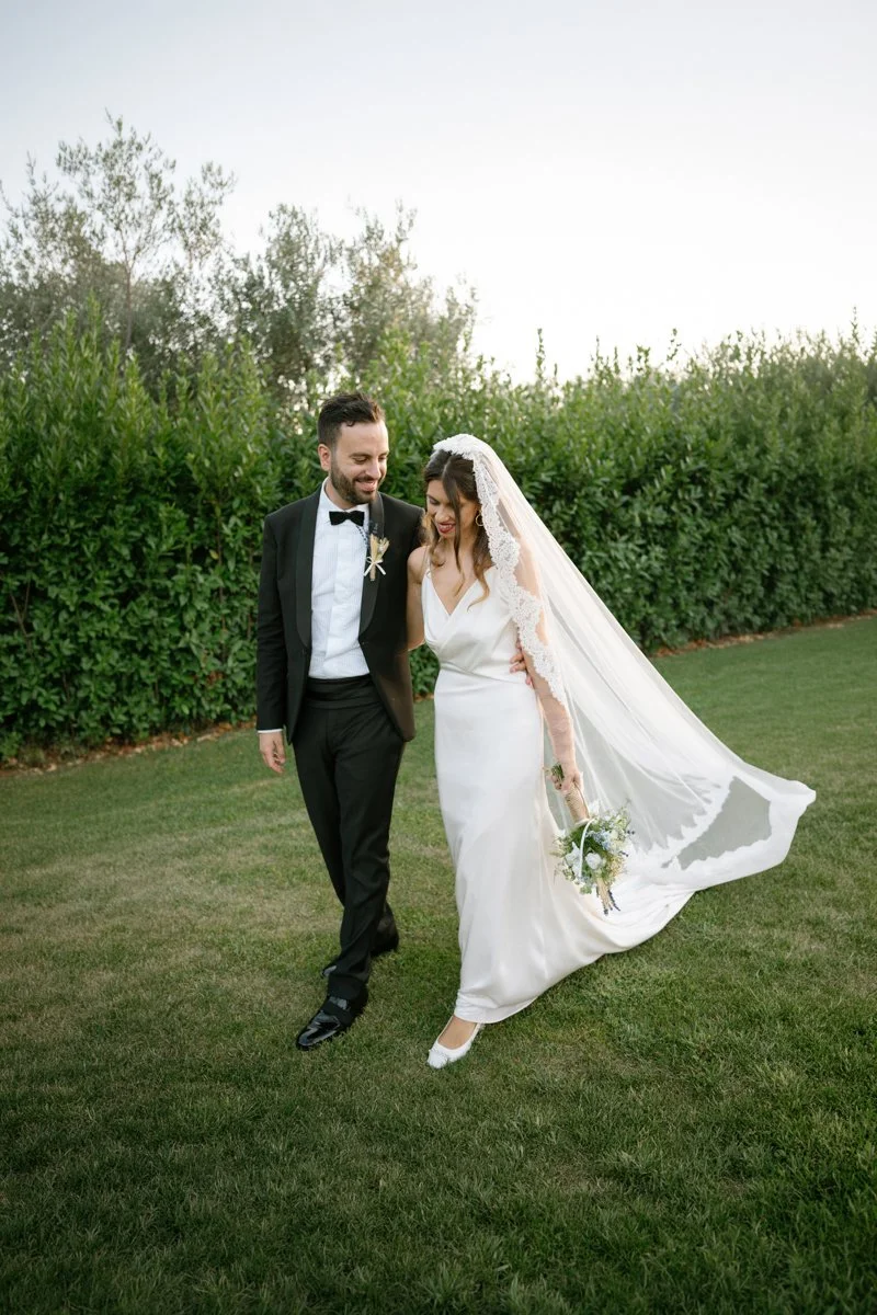 A newlywed couple walking on a grassy lawn, smiling and holding each other, with the bride holding a bouquet, against a background of green bushes and trees.