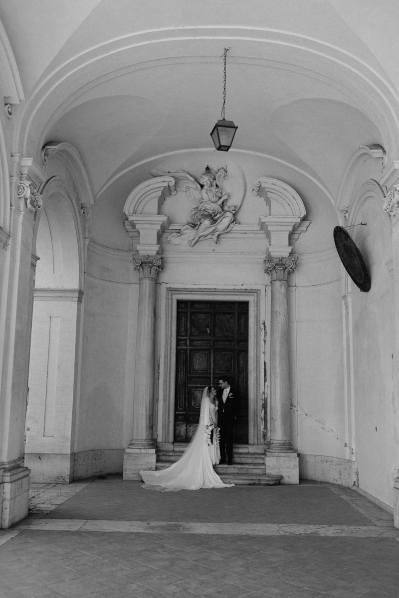 A bride and groom standing close together inside a historic building with high ceilings, ornate columns, and a decorative sculpture of an angel or mythological figure above them. The bride is wearing a long train wedding dress, holding a bouquet, and