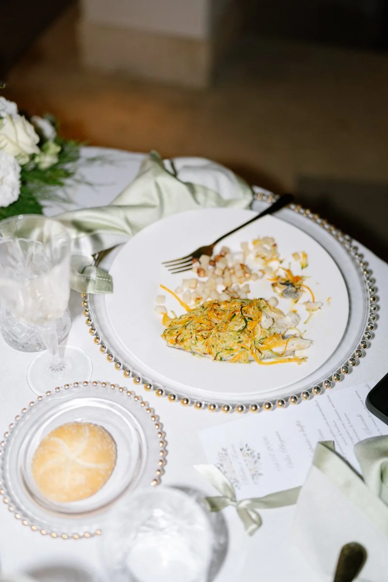A partially eaten plate of food with shredded vegetables and a side of diced food, a fork, and a glass of water on a decorated table with a menu and a floral arrangement.
