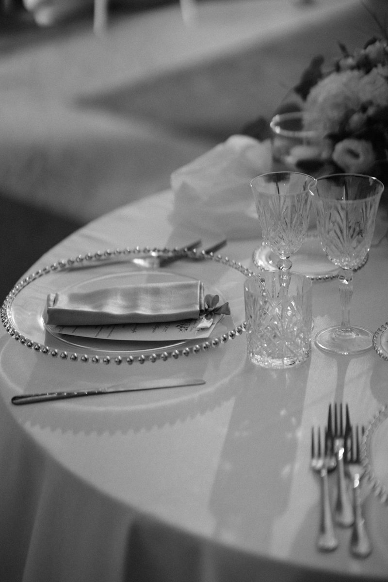 A formal dining table setting with crystal glasses, silverware, a napkin with a bow, and floral arrangements in the background, all on a round table with a white tablecloth.