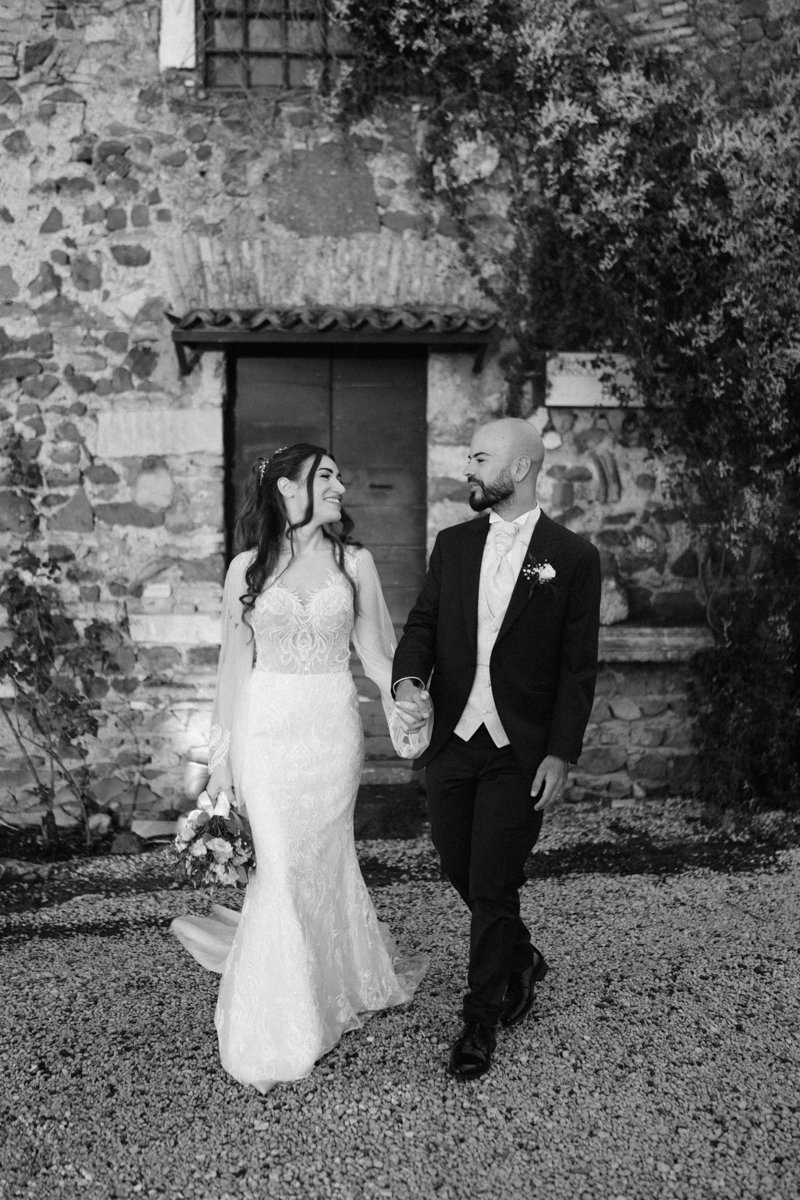 A black and white photo of a bride and groom walking hand in hand outside a rustic stone building with a door and shingles, surrounded by trees and foliage, during their wedding.