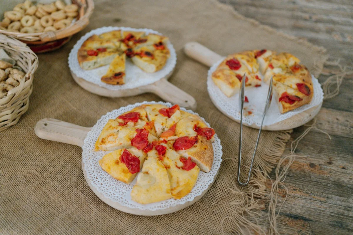 Three small pizza slices with cheese and tomato toppings on lace-edged paper plates on wooden serving paddles, with tongs, on a rustic wooden table with burlap cloth, alongside baskets of snacks.