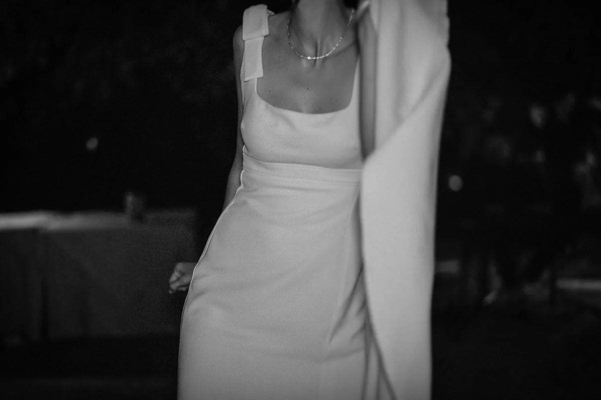 Black and white photo of a woman wearing a sleeveless dress with a square neckline and a pearl necklace, outdoors at night.