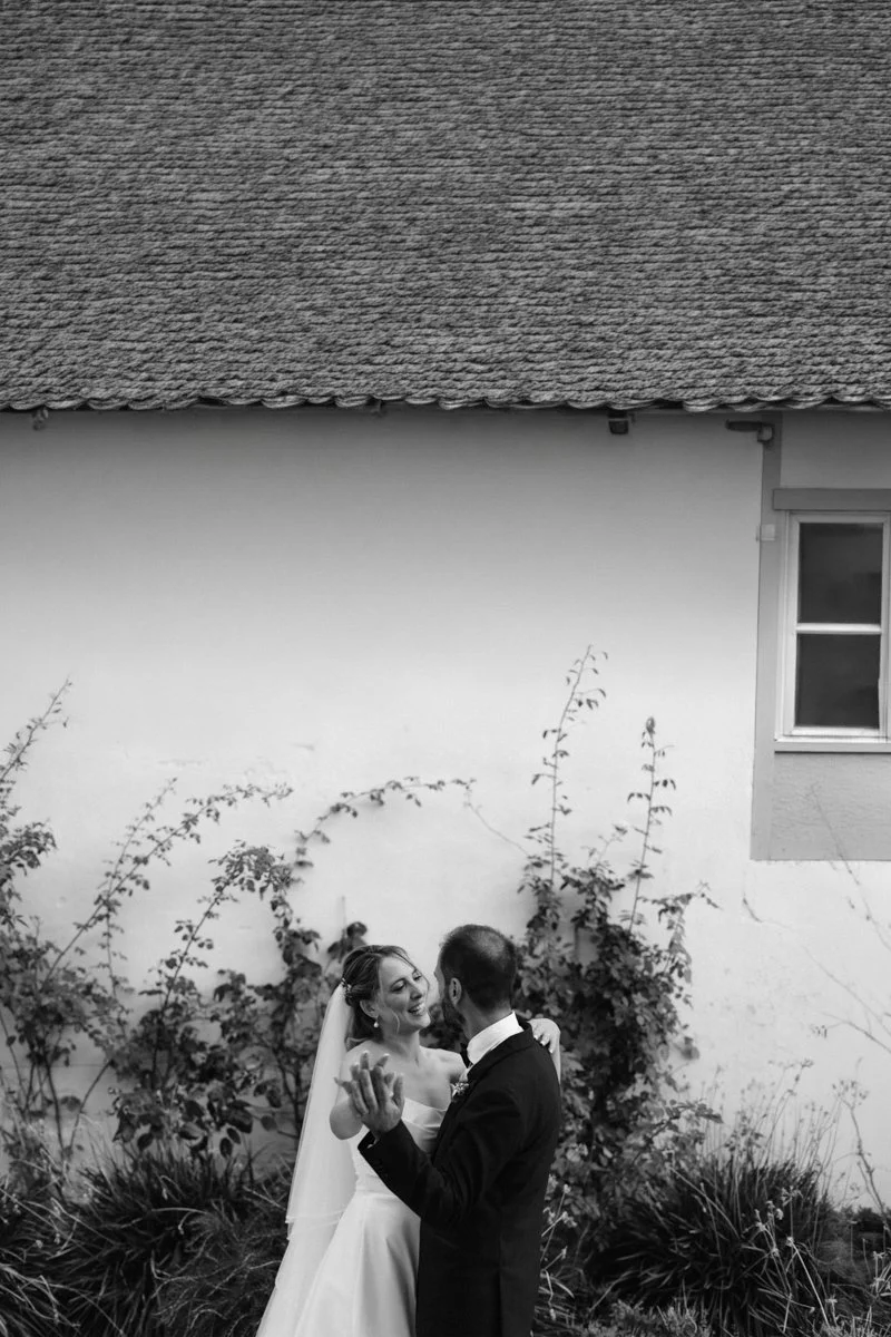 A bride and groom dancing outdoors against a white wall with climbing plants and a window in the background, in black and white.