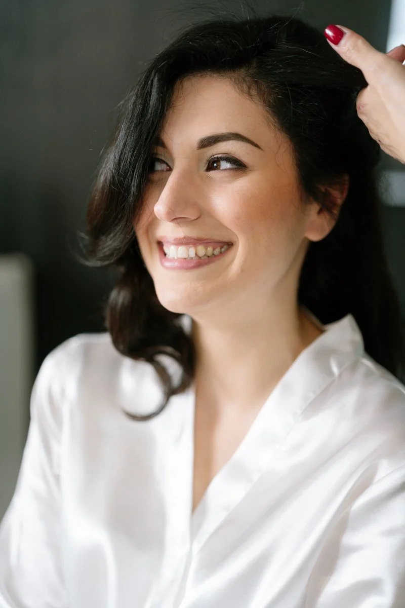 A woman with dark hair and a white satin robe smiling as someone styles her hair.