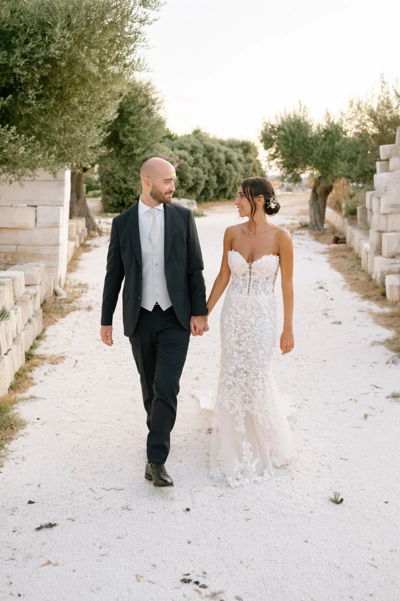 A bride and groom walking hand in hand outdoors on a sandy path lined with trees and stone structures, dressed in wedding attire.
