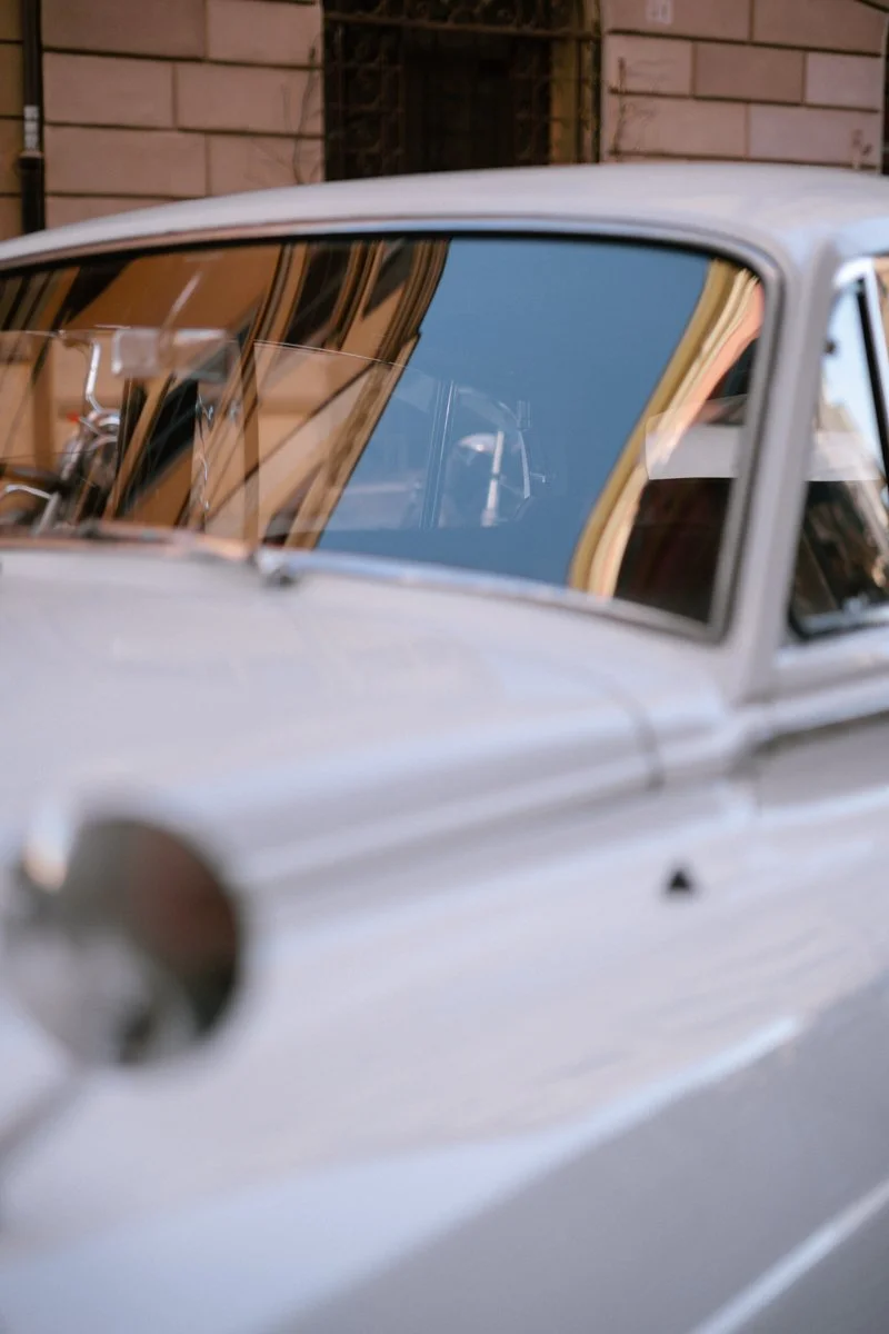Close-up of a vintage white car with a reflective windshield, parked on a city street with buildings reflected on the glass.