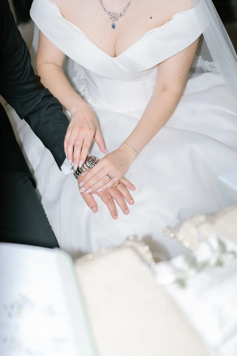 Close-up of a bride and groom holding hands, with wedding rings, on her wedding dress and jewelry visible.