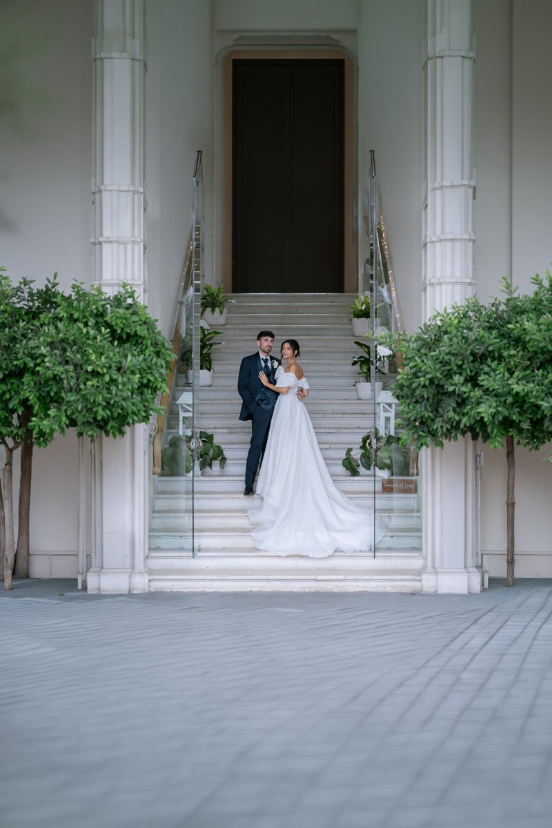 A bride and groom standing on white marble stairs in front of a large dark door, surrounded by greenery and potted plants, possibly outside a wedding venue or church.