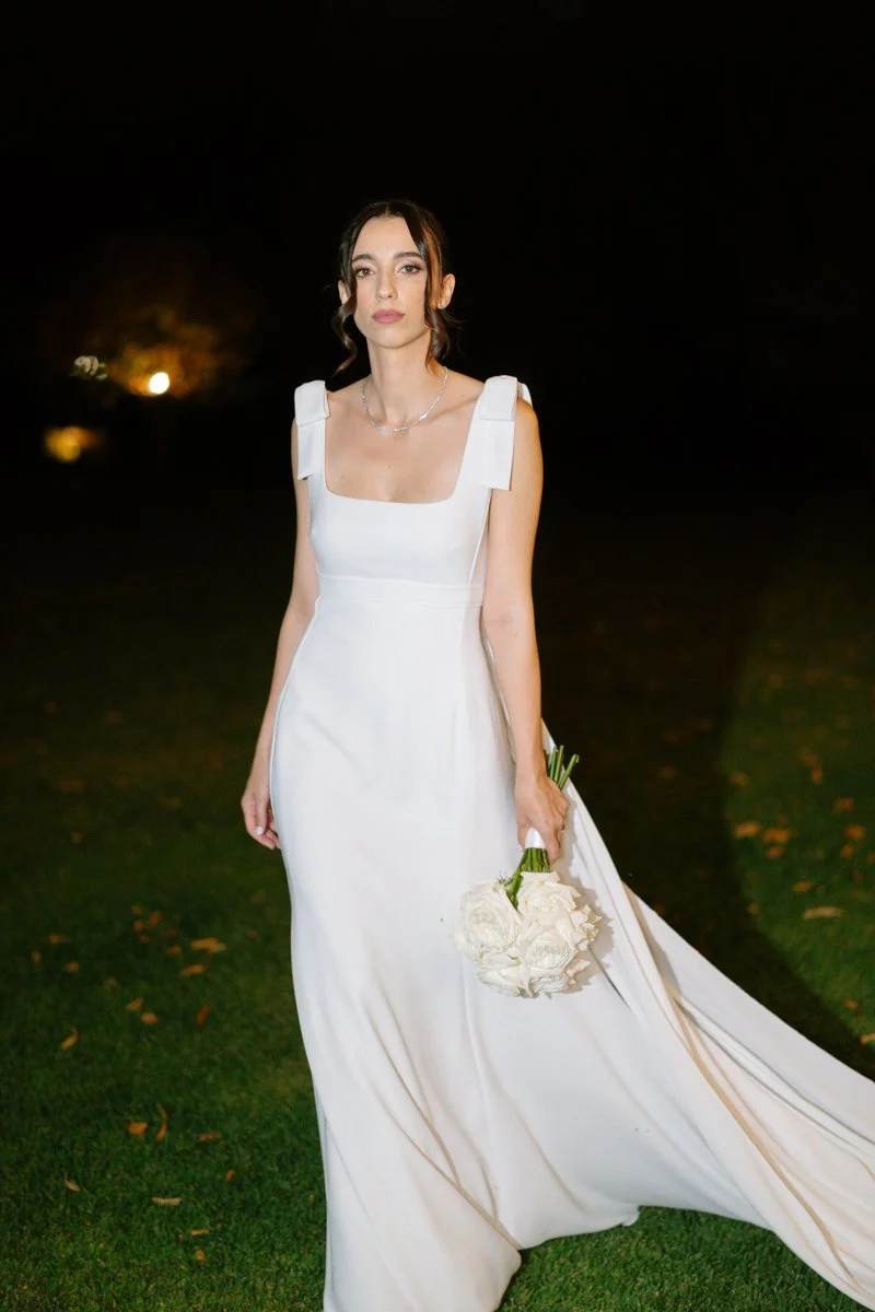 A woman in a white wedding dress holding a bouquet of white flowers at night outside.