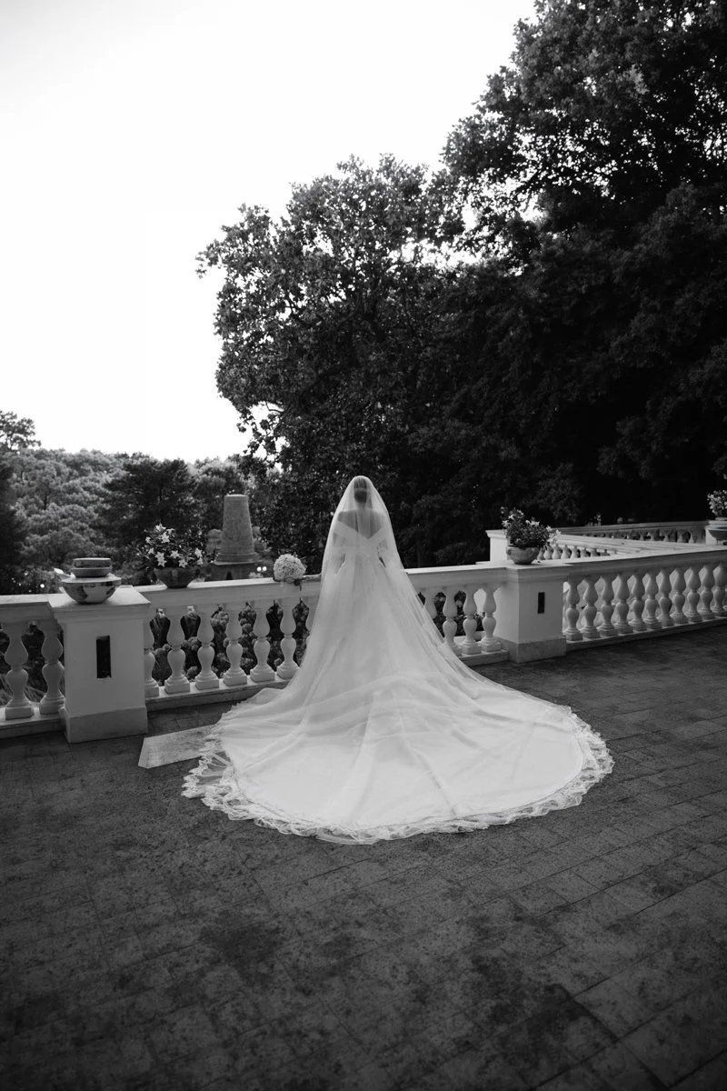 A bride in a wedding gown with a long train and veil stands on a balcony with a scenic outdoor view of trees.