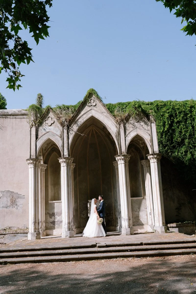A bride and groom standing face to face at an old, Gothic-style stone chapel with tall arches and columns during daytime.