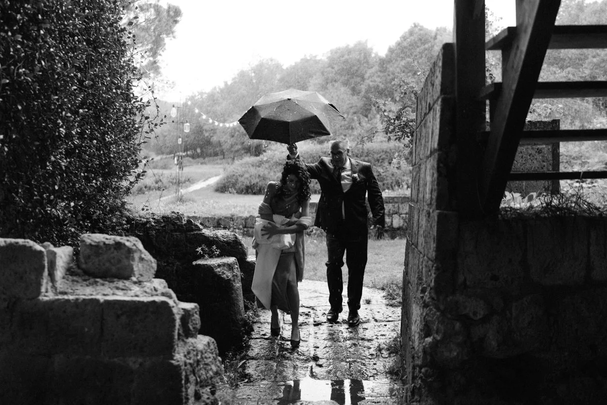 A man in a suit and a woman in a dress walk together under an umbrella on a rainy day outdoors, walking past stone steps and a stone wall, with trees and a pathway in the background.