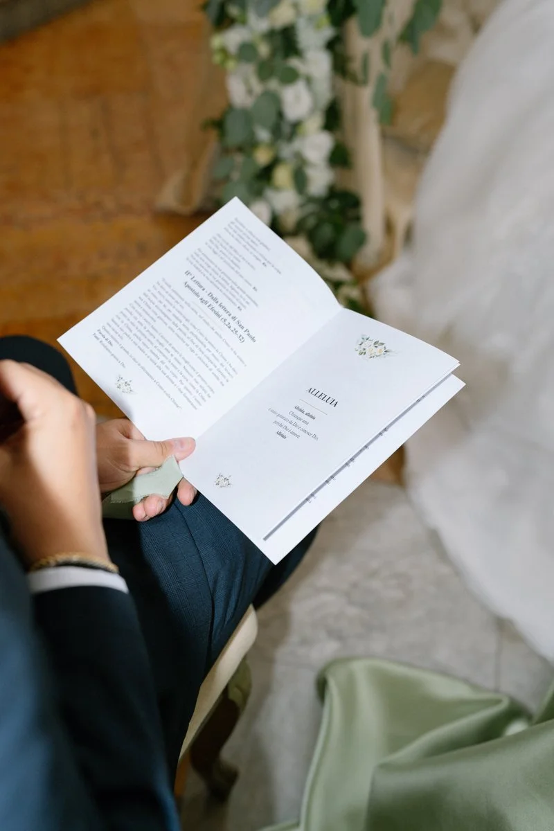 Person reading a wedding program or booklet at a wedding ceremony, with white flowers and greenery in the background.