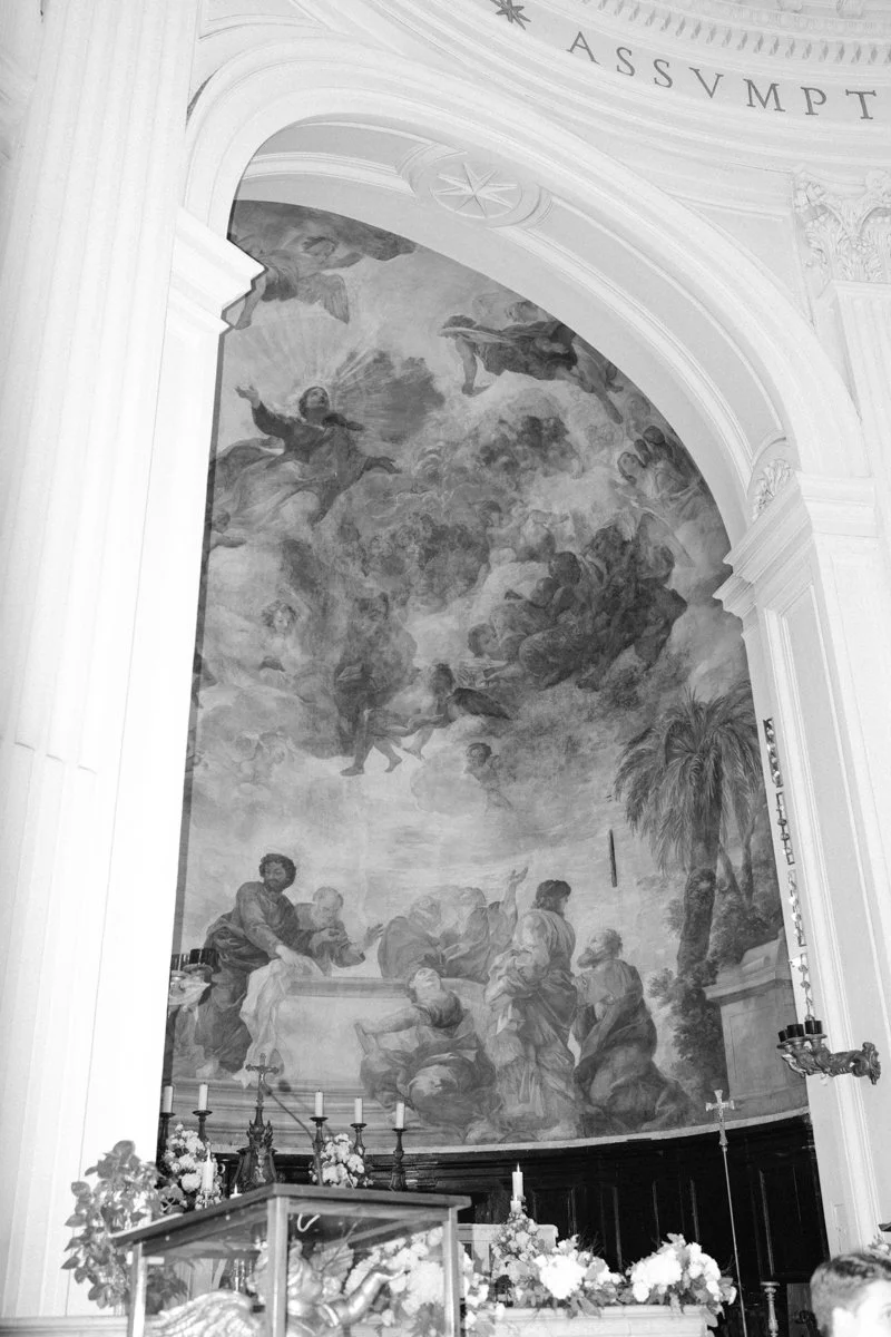A large religious mural inside a church depicting angels, clouds, and biblical figures with some flowers and candles near the altar.