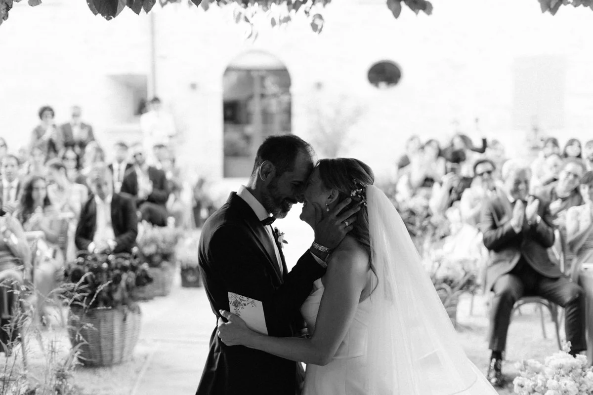 A black and white photo of a wedding ceremony. The bride and groom are touching foreheads and holding each other, with guests applauding in the background.
