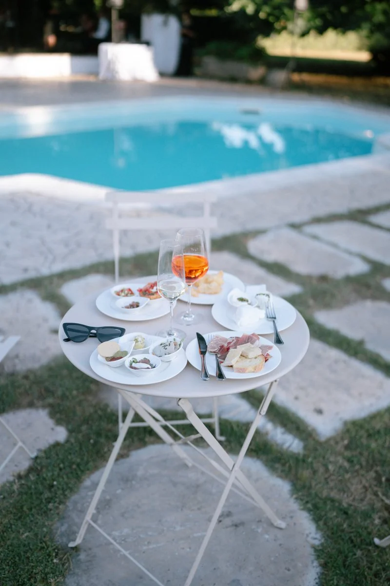 A small round table with plates of food, glasses of drinks, and sunglasses set outside near a swimming pool, with chairs and greenery in the background.