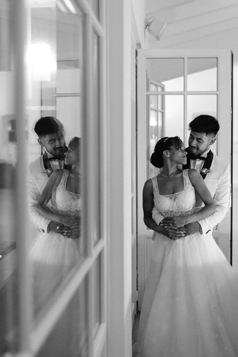 A black-and-white photo of a couple in wedding attire, standing close together and looking affectionately at each other, with their reflection visible in a mirror.