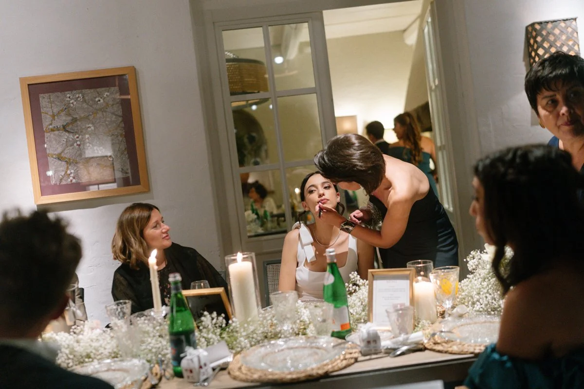 A woman is applying makeup to a seated woman at a formal dinner party with several other guests. The dining table is decorated with candles, flowers, and drinks.
